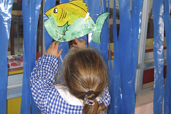 La imagen muestra a una niña pegando un dibujo de un pez en un fondo de plástico azul