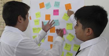 La imagen muestra a dos estudiantes debatiendo frente a una pizarra con post it de diferentes colores