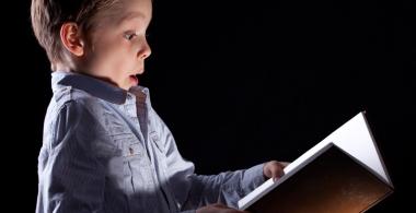 La imagen muestra a un niño en fondo negro mirando un libro abierto entre sus manos con cara de asombro