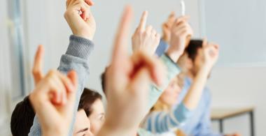 La imagen muestra estudiantes levantando la mano