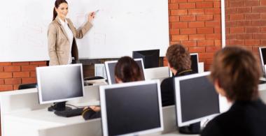 La imagen muestra estudiantes en sala de computadores con profesora entregando contenido