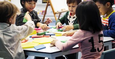 La imagen muestra un grupo de niños y niñas en una mesa interactuando con diversos objetos de tamaños y colores diferentes